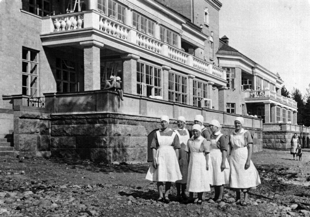 Kuusi valkoiseen esiliinaan ja päähuiviin pukeutunutta naista ison kivirakennuksen edessä. Rakennuksessa näyttävät kuistit, jonka kaiteella istuu muutama lasi. Kuva on mustavalkoinen.