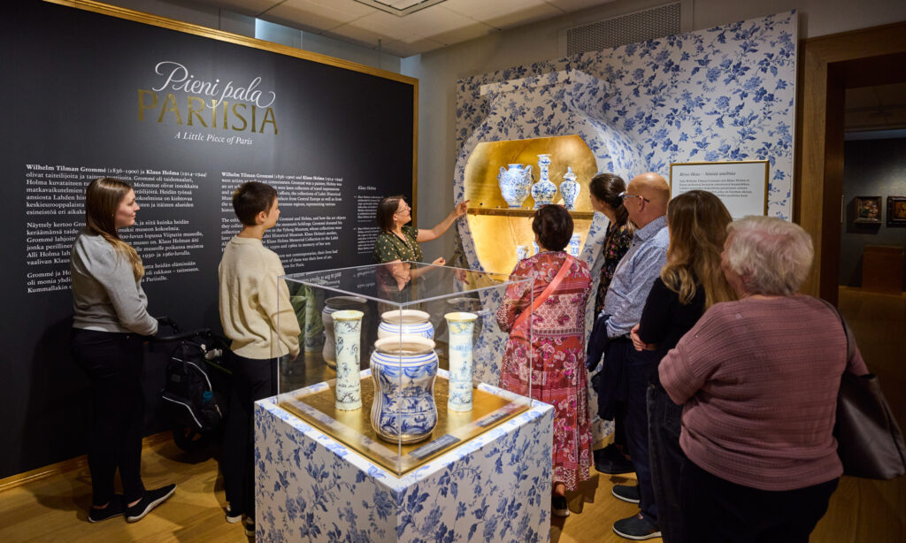 Museokävijöitä kokoontuneena kuuntelemaan opasta, joka osoittaa isoa maljakon muotoista vitriiniä, jonka sisällä on keramiikkaa. Ryhmän edessä keskilattialla myös keramiikkavitriini.