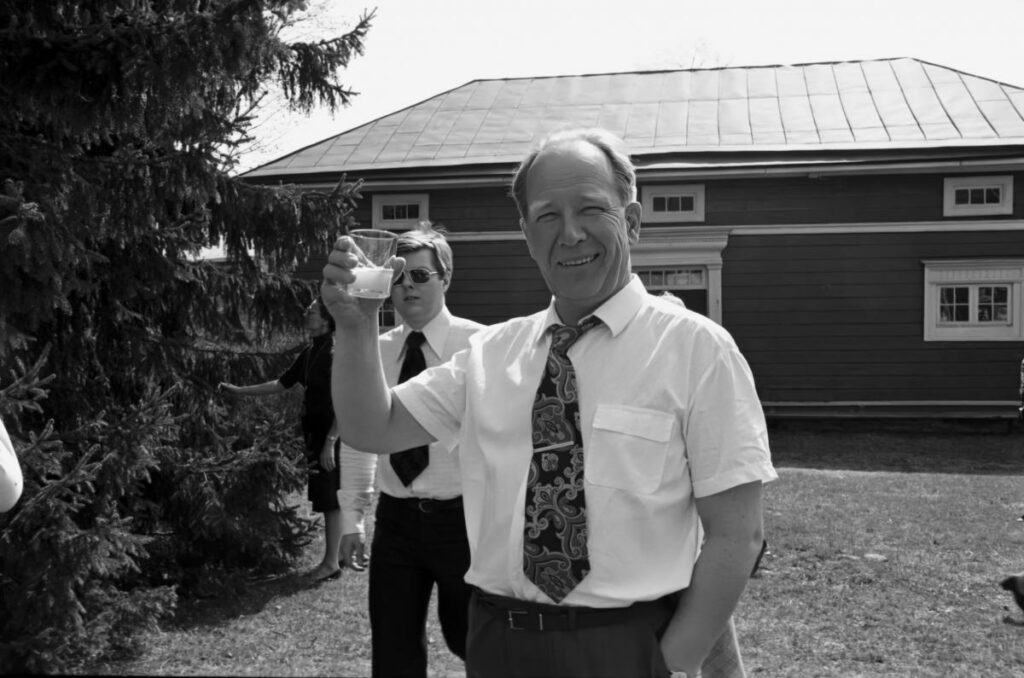 Valkoiseen kauluspaitaan ja solmioon pukeutunut mies kohottaa puolillaan olevaa lasia hymyillen. Kuva on mustavalkoinen.